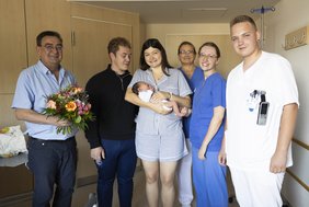 Eine Gruppe aus Arzt, Hebamme und Pflegefachkraft steht gemeinsam mit dem stellvertretenden Geschäftsführer bei den Eltern, die das Baby im Arm halten. Sie beglückwünschen diese zur Geburt. 