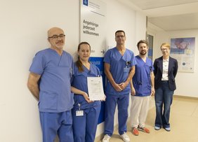Nebeneinander steht das Leitungsteam der Intensivstation aus Pflegekräften und Ärzten mit der Geschäftsführerin des Krankenhauses. Sie halten ein Zertifikat ins Bild. Auf dem Zertifikat steht, dass das Krankenhaus erneut zertifiziert wurde.