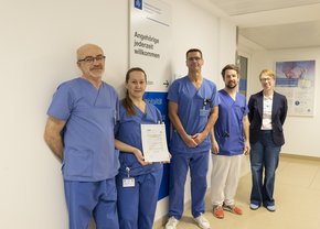 Nebeneinander steht das Leitungsteam der Intensivstation aus Pflegekräften und Ärzten mit der Geschäftsführerin des Krankenhauses. Sie halten ein Zertifikat ins Bild. Auf dem Zertifikat steht, dass das Krankenhaus erneut zertifiziert wurde.